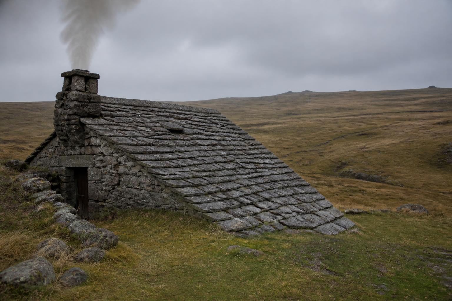 Buron en pierre de lauze à moitié enterré dans une pâture de l'Aubrac, de la fumée s'échappe de la cheminée, ciel d'automne gris pâle