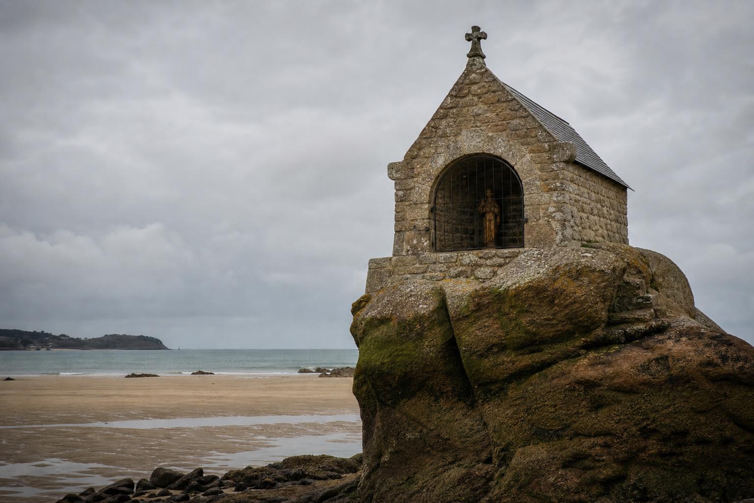 L'oratoire de Saint-Guirec sur son rocher à marée basse, sous un ciel gris d'hiver, la statue de bois visible derrière la grille, l'anse déserte tout autour