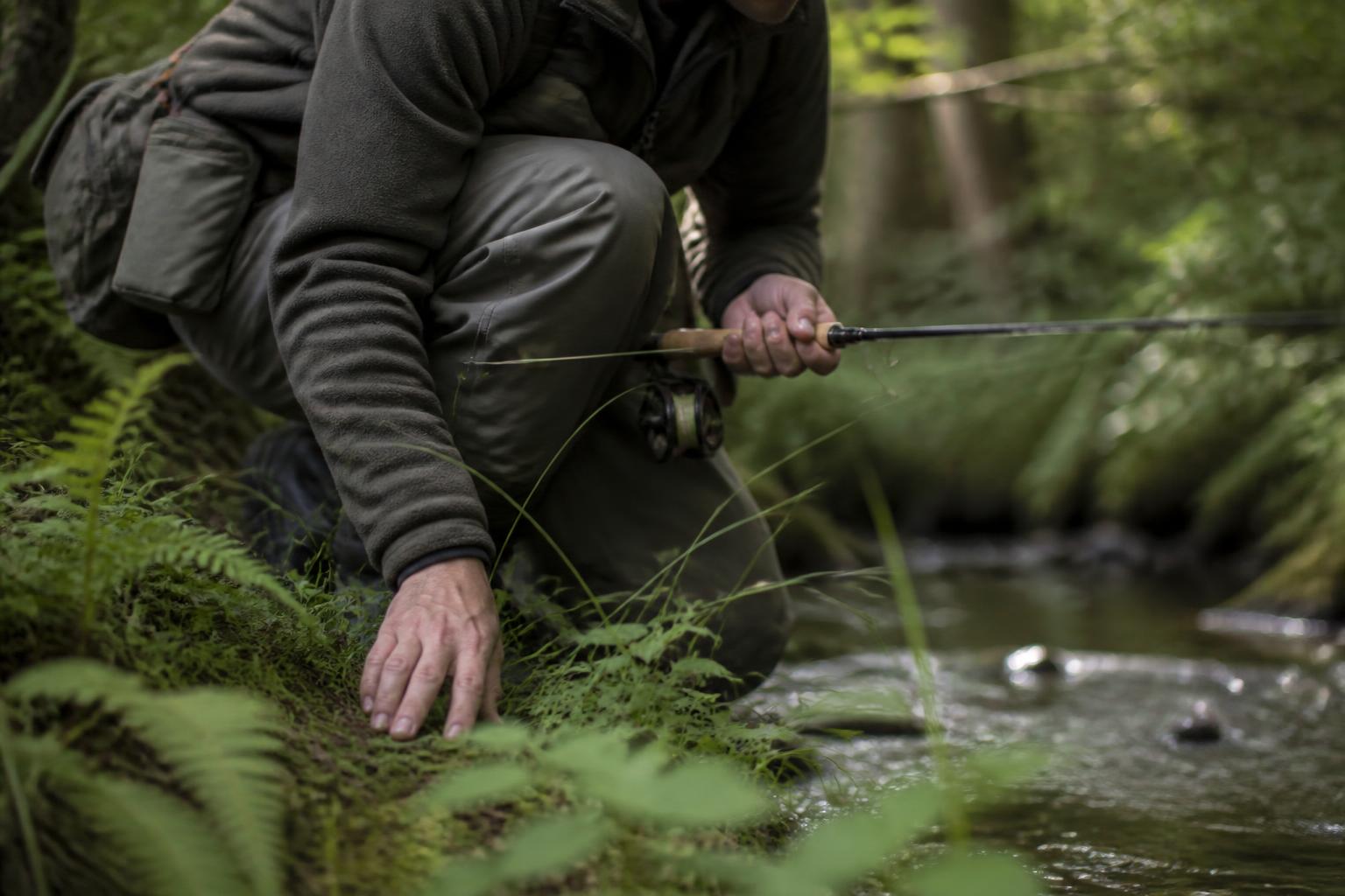 Pêcheur accroupi au bord d'une petite rivière, canne en main, silhouette basse pour ne pas être repéré par la truite