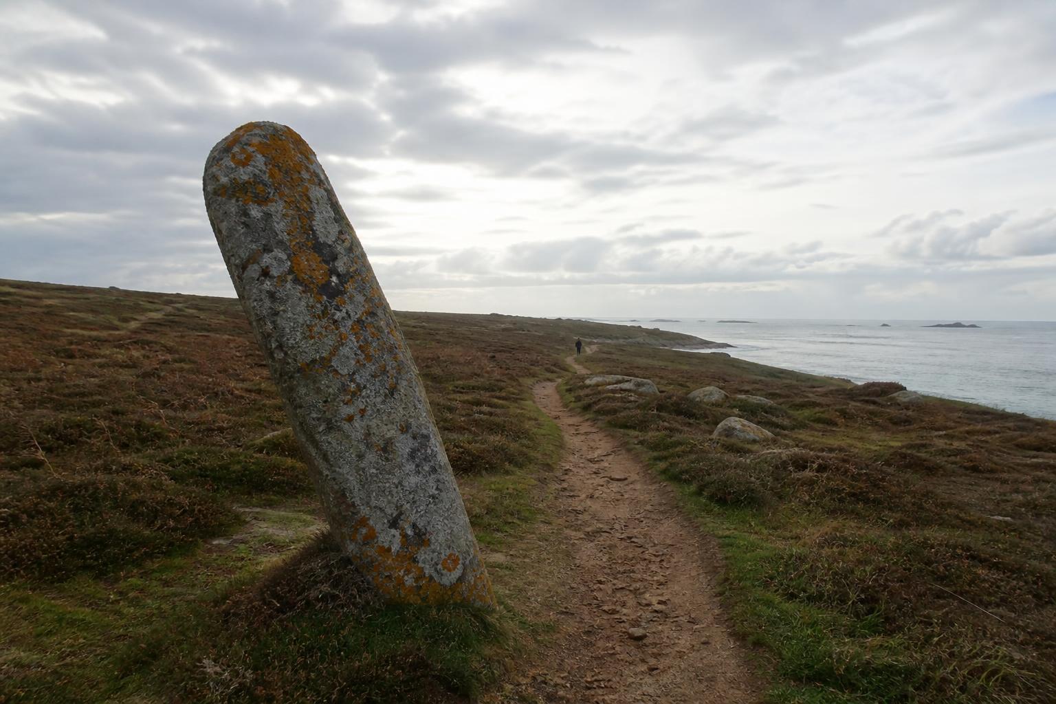 Le sentier côtier de l'Île-Grande sous un ciel de février, un menhir couvert de lichen orange au premier plan, la mer grise au loin, une silhouette de promeneur sur le chemin